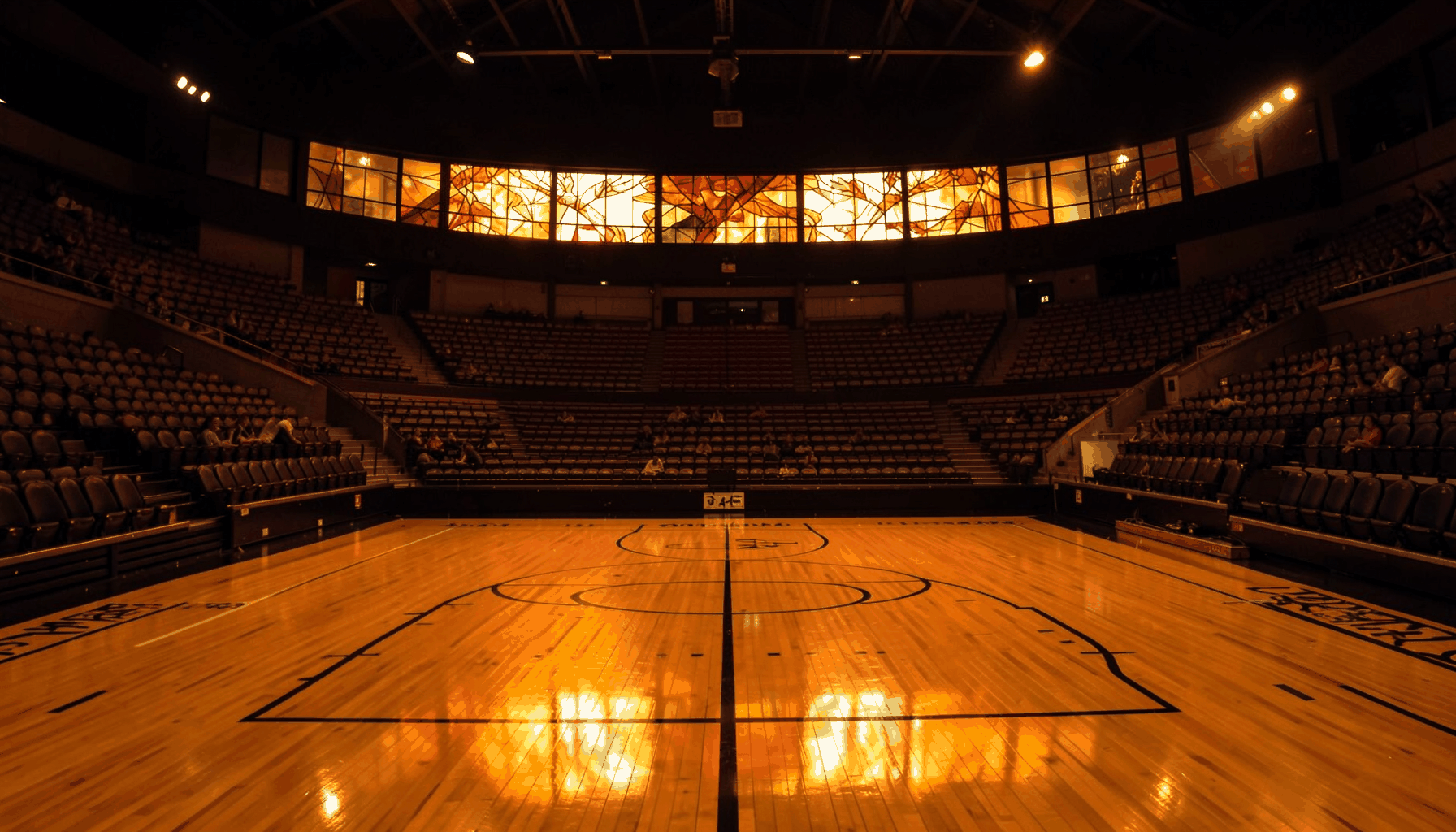 A warm, atmospheric photograph of a basketball court seen from the stands, polished wood gleaming under amber arena lights