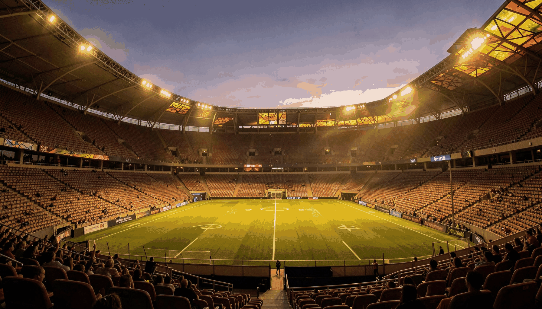 Wide atmospheric photograph of a stadium at dusk, warm amber and tan tones washing over empty seats, the field glowing under soft evening light